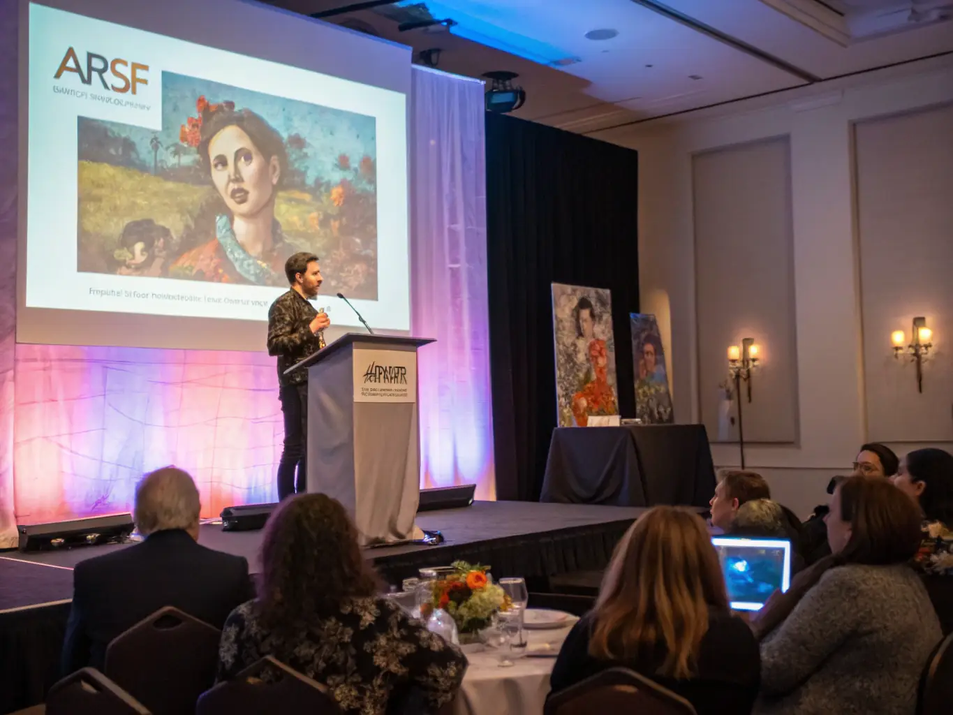 A photograph of an artist giving a talk about their work, with an attentive audience listening in a gallery setting during the Lurcy Les Arts exhibition.